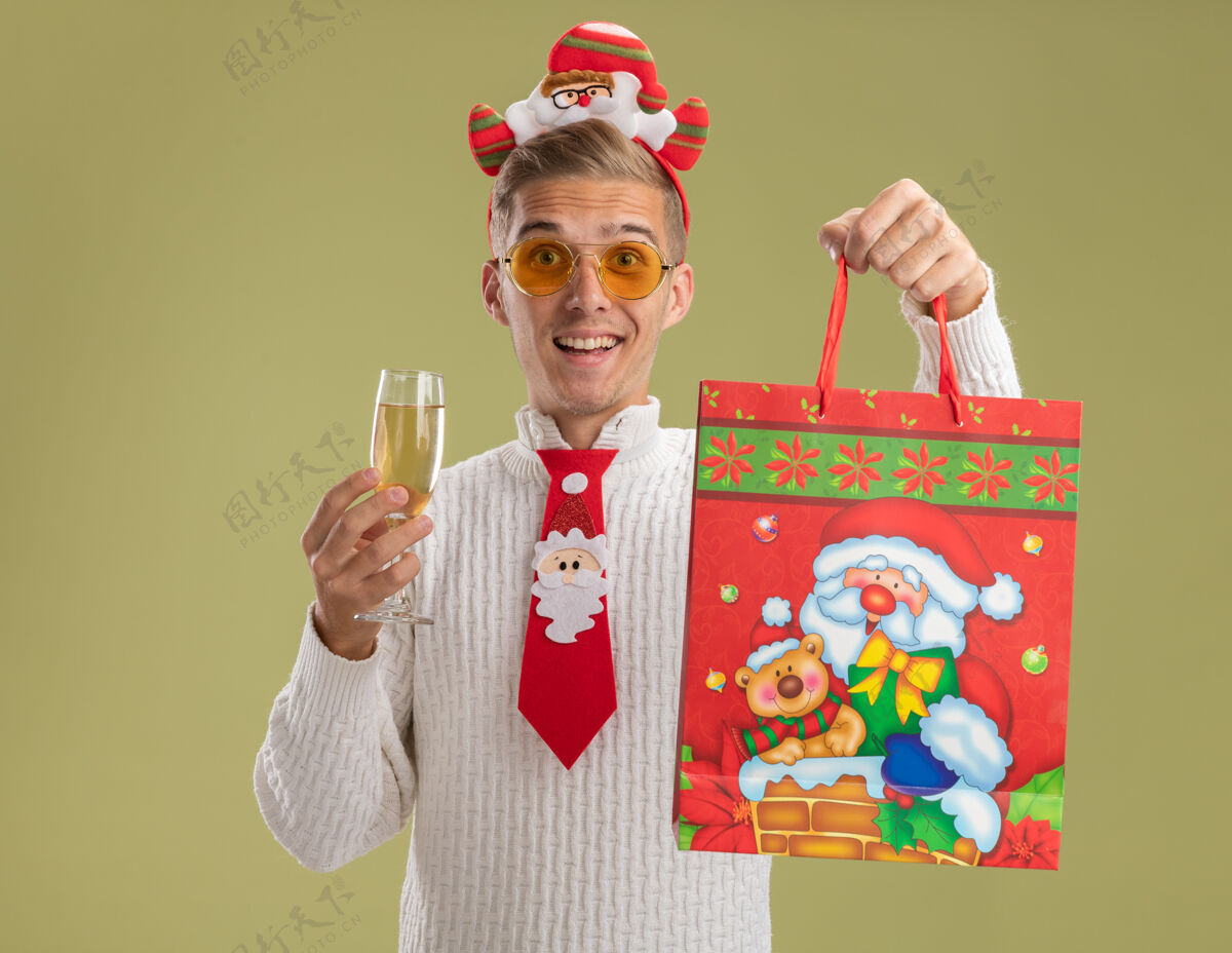 圣诞老人令人印象深刻的年轻帅哥戴着圣诞老人的头带和领带拿着一杯香槟和圣诞礼包隔离在橄榄绿的墙上印象深刻领带年轻
