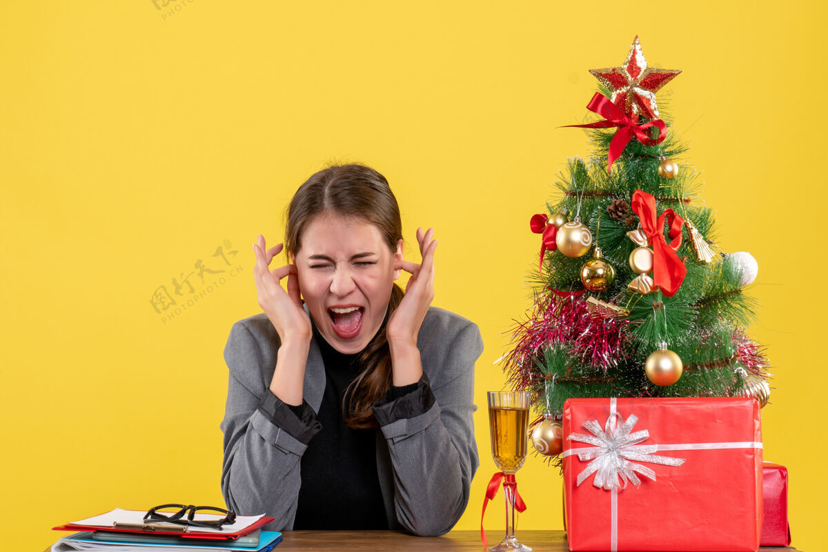 女孩坐在书桌旁的女孩大声喊道 用手指捂住耳朵 圣诞树和礼物鸡尾酒桌子圣诞节生活