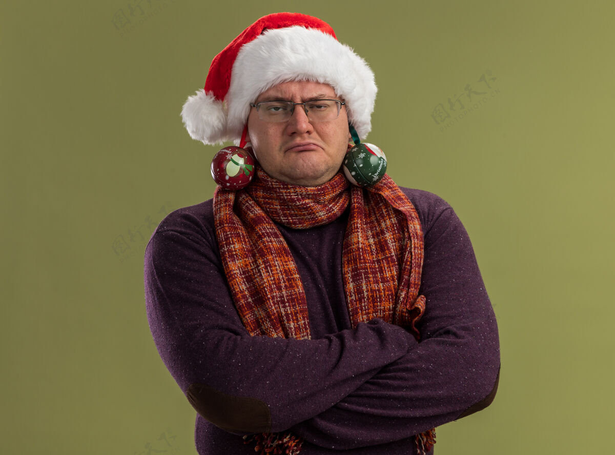 围巾自信的成年男子戴着眼镜 戴着圣诞帽 脖子上围着围巾 站着 闭着身子看着相机 耳朵上挂着圣诞饰品 背景是橄榄绿的脖子圣诞老人饰品