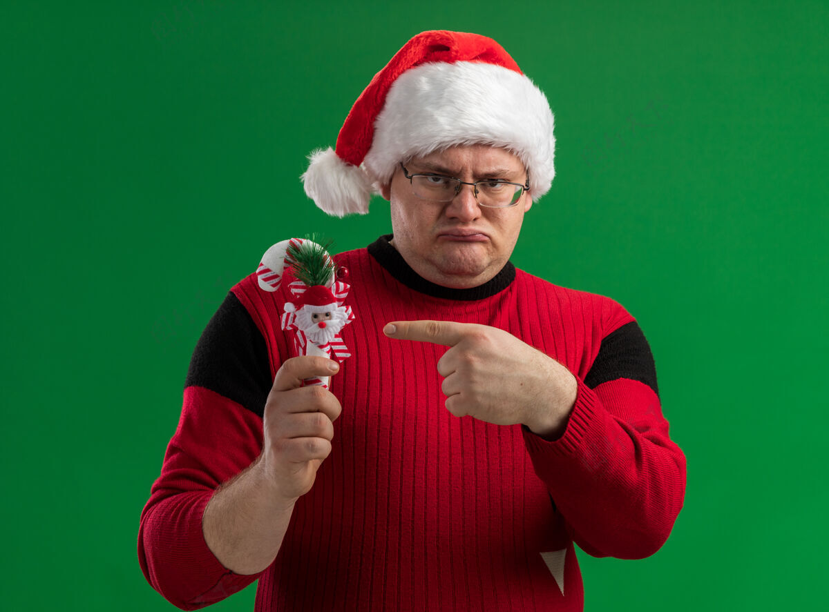 成人愁眉不展的成年男子戴着眼镜 拿着圣诞老人的帽子 指着糖果藤饰品 看着隔离在绿色背景上的相机糖果拿着帽子