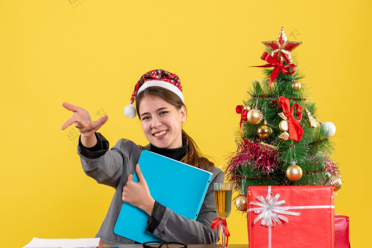 桌子前视图微笑着戴圣诞帽的女孩坐在圣诞树和礼物鸡尾酒桌旁帽子成人礼物