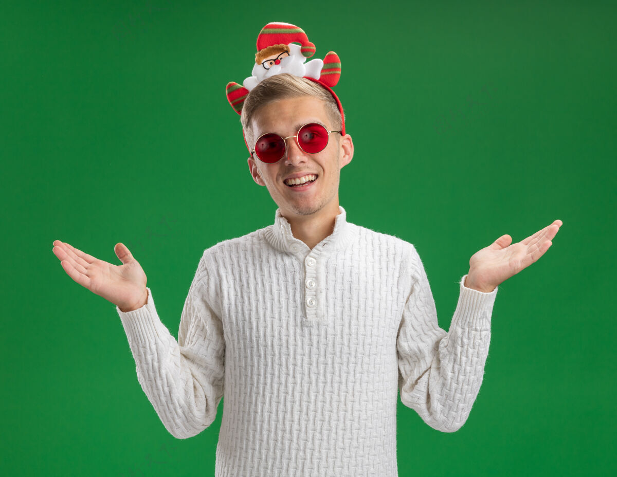 圣诞老人快乐的年轻帅哥戴着圣诞老人的头带 戴着眼镜 两手空空的 孤立在绿色的墙上空虚穿着圣诞老人