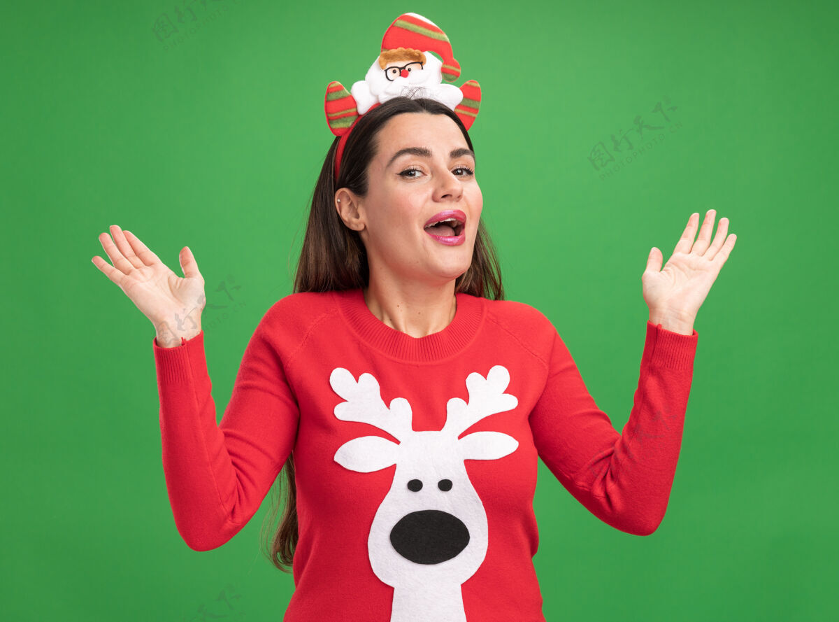 手惊讶的年轻漂亮女孩穿着圣诞毛衣与圣诞发箍展开双手孤立的绿色背景头发蔓延女孩