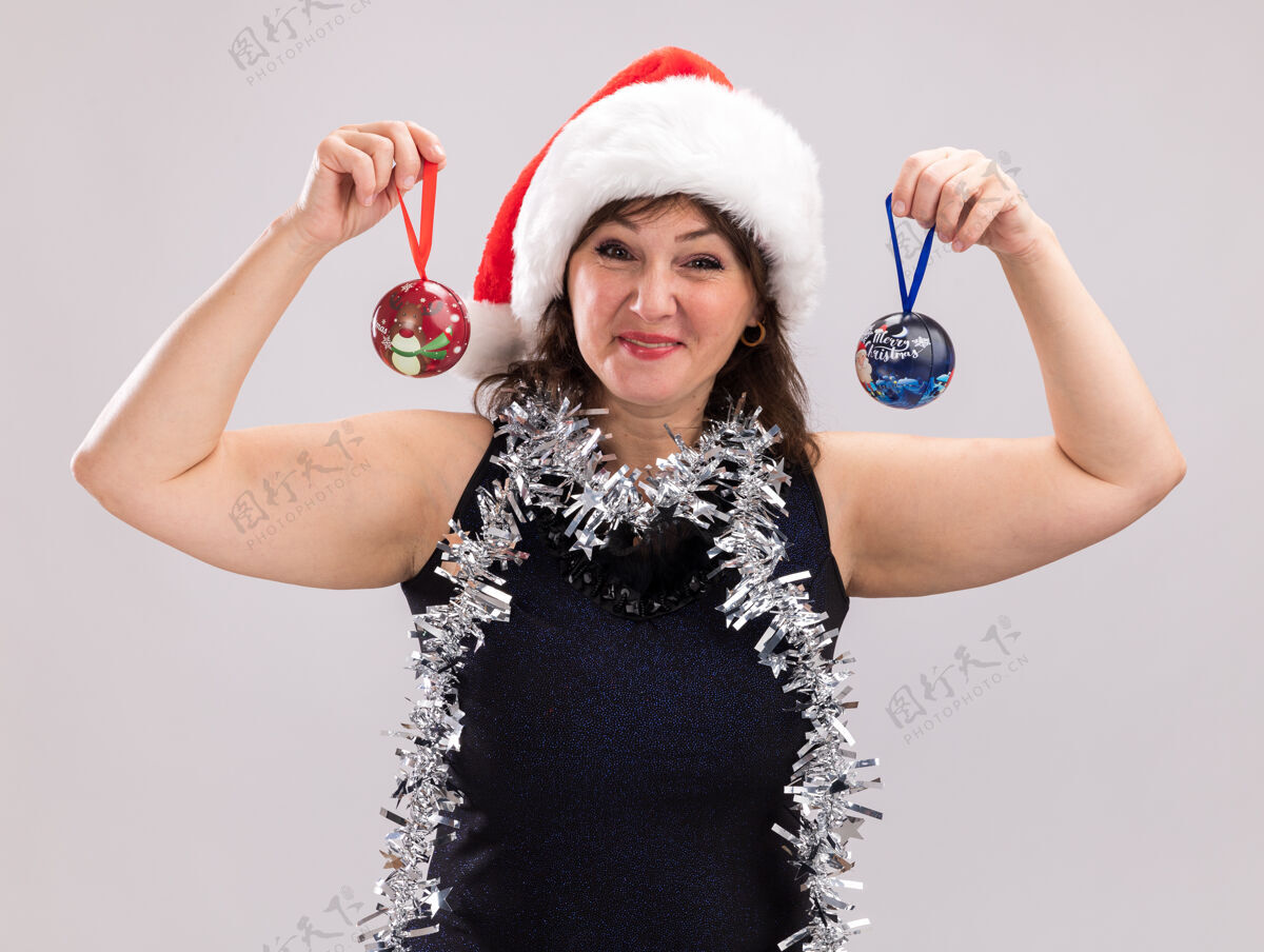 花环高兴的中年妇女戴着圣诞帽 脖子上戴着金箔花环 头上拿着圣诞饰品 看着隔离在白色背景上的相机中年圣诞节脖子