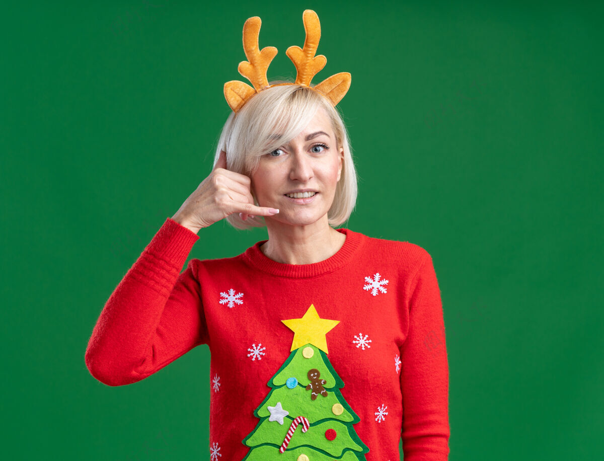 看微笑的中年金发女人戴着圣诞驯鹿鹿角头带和圣诞毛衣看着相机做着呼叫手势 在绿色背景上与复制空间隔离鹿角中年电话
