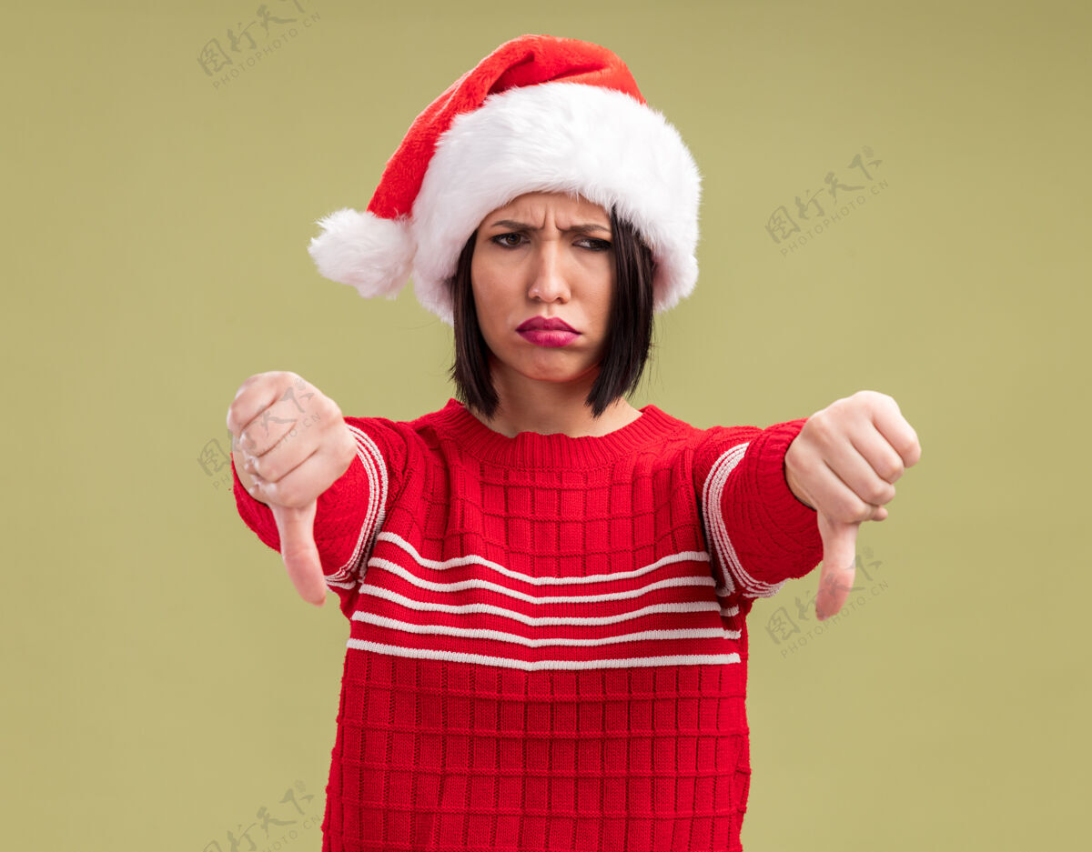 向下愁眉苦脸的年轻女孩戴着圣诞帽 看着一边竖起大拇指 孤立地站在橄榄绿的背景上拇指侧圣诞老人