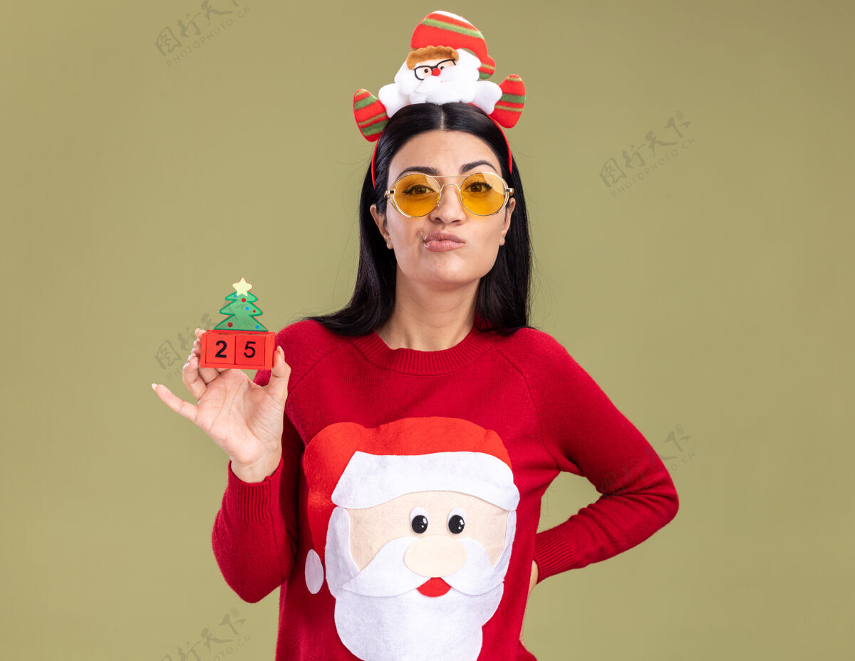 自信自信的年轻白人女孩戴着圣诞老人的头带和带眼镜的毛衣拿着圣诞树玩具手放在腰上看着橄榄绿背景上的相机圣诞圣诞老人相机