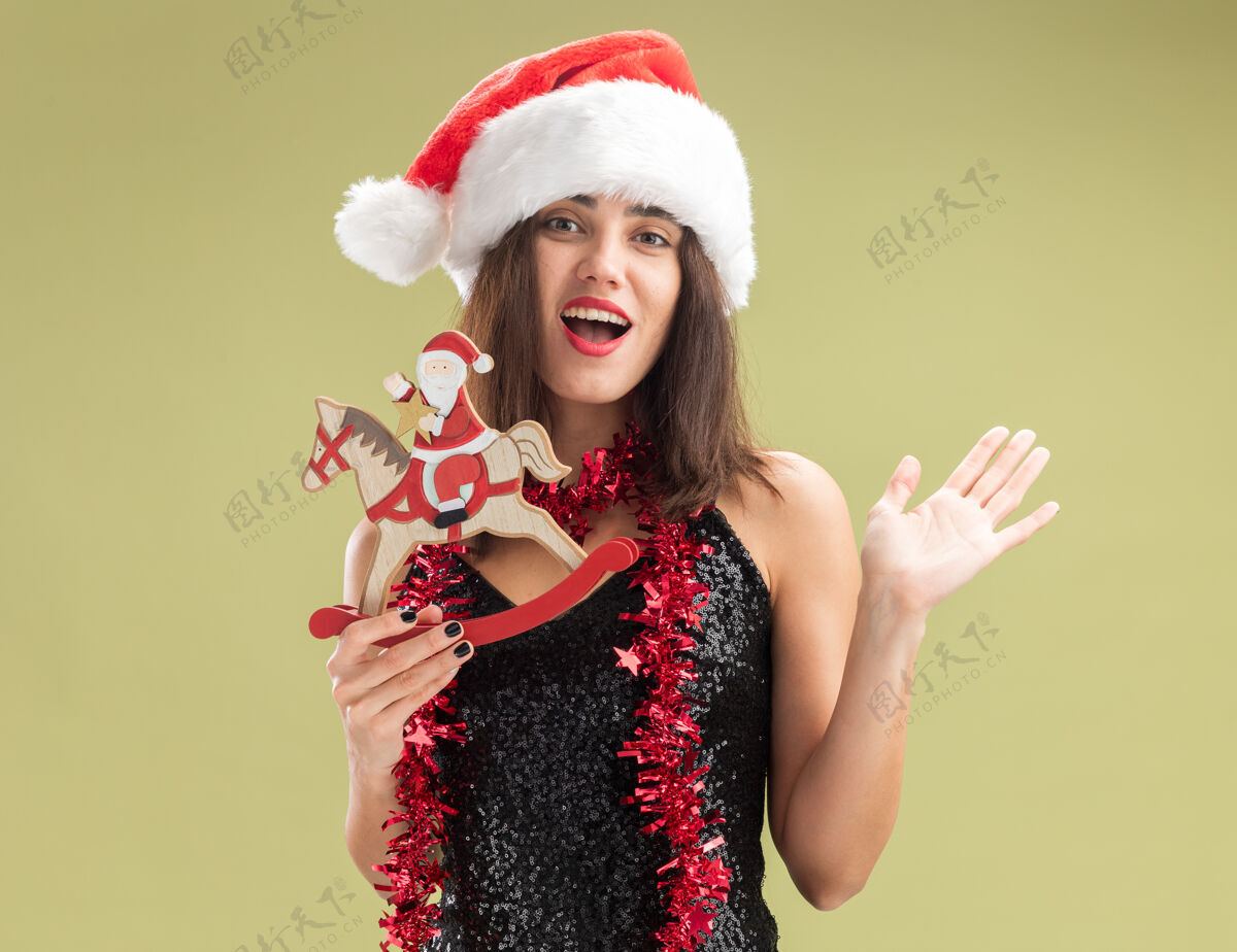 脖子惊讶的年轻漂亮的女孩戴着圣诞帽 脖子上戴着花环 手里拿着圣诞玩具 在橄榄绿的背景上孤立着橄榄抱着惊讶