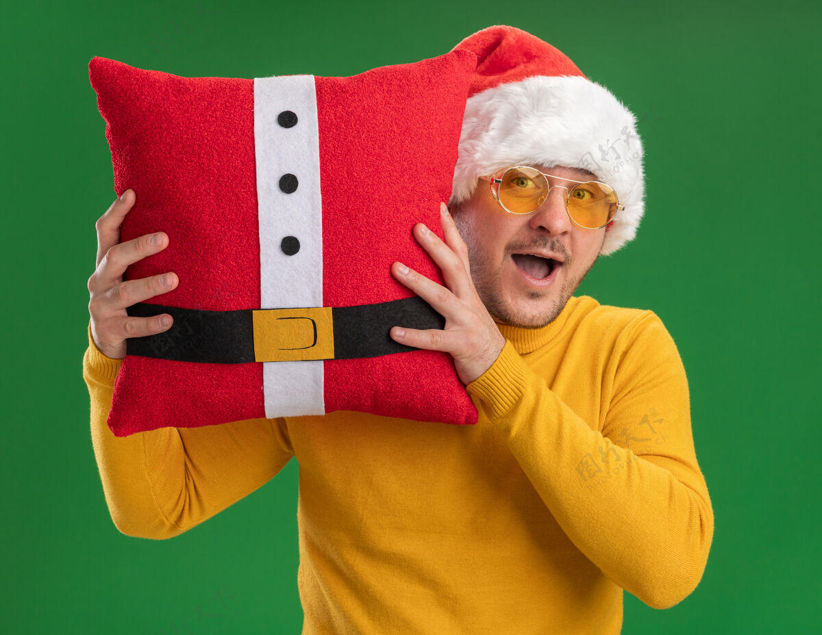 眼镜快乐的年轻人穿着黄色高领毛衣 戴着圣诞帽 戴着眼镜 抱着红色滑稽的枕头 站在绿色的背景下惊讶地看着相机搞笑圣诞老人年轻
