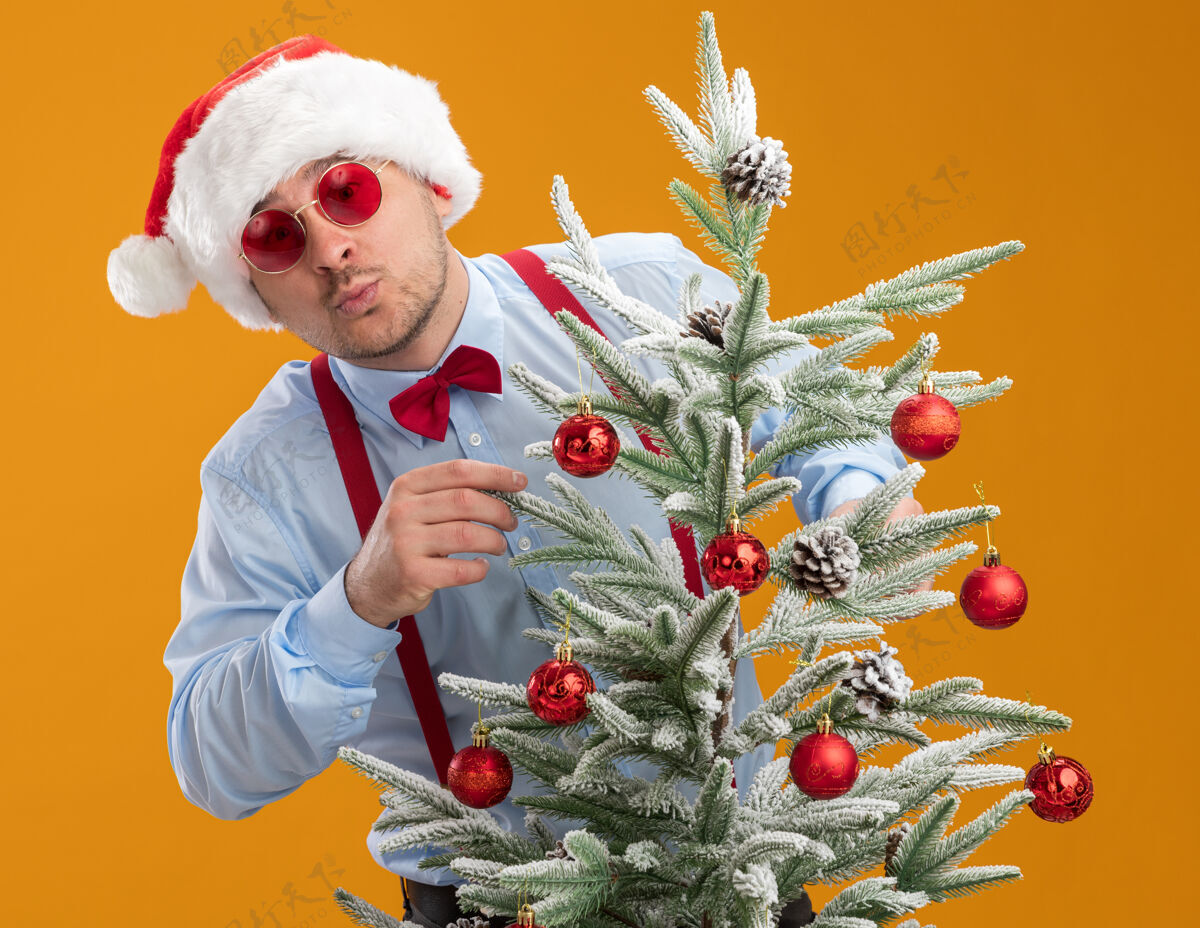 圣诞节戴着圣诞帽 戴着红眼镜 系着吊带领结的年轻人站在圣诞树后面 高兴而惊奇地把玩具挂在橙色背景的树上帽子吊带站立