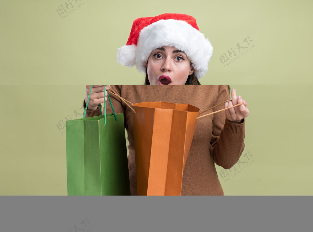 穿着年轻漂亮的女孩戴着圣诞帽拿着礼品袋孤立在橄榄绿的墙上关心圣诞节帽子
