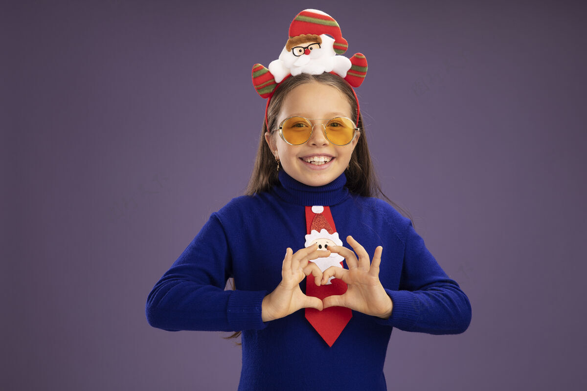 心小女孩穿着蓝色高领毛衣 打着红色领带 头上戴着有趣的圣诞戒指 看着相机 开心地微笑着 做着心形手势 手指站在紫色的背景上愉快手指边缘