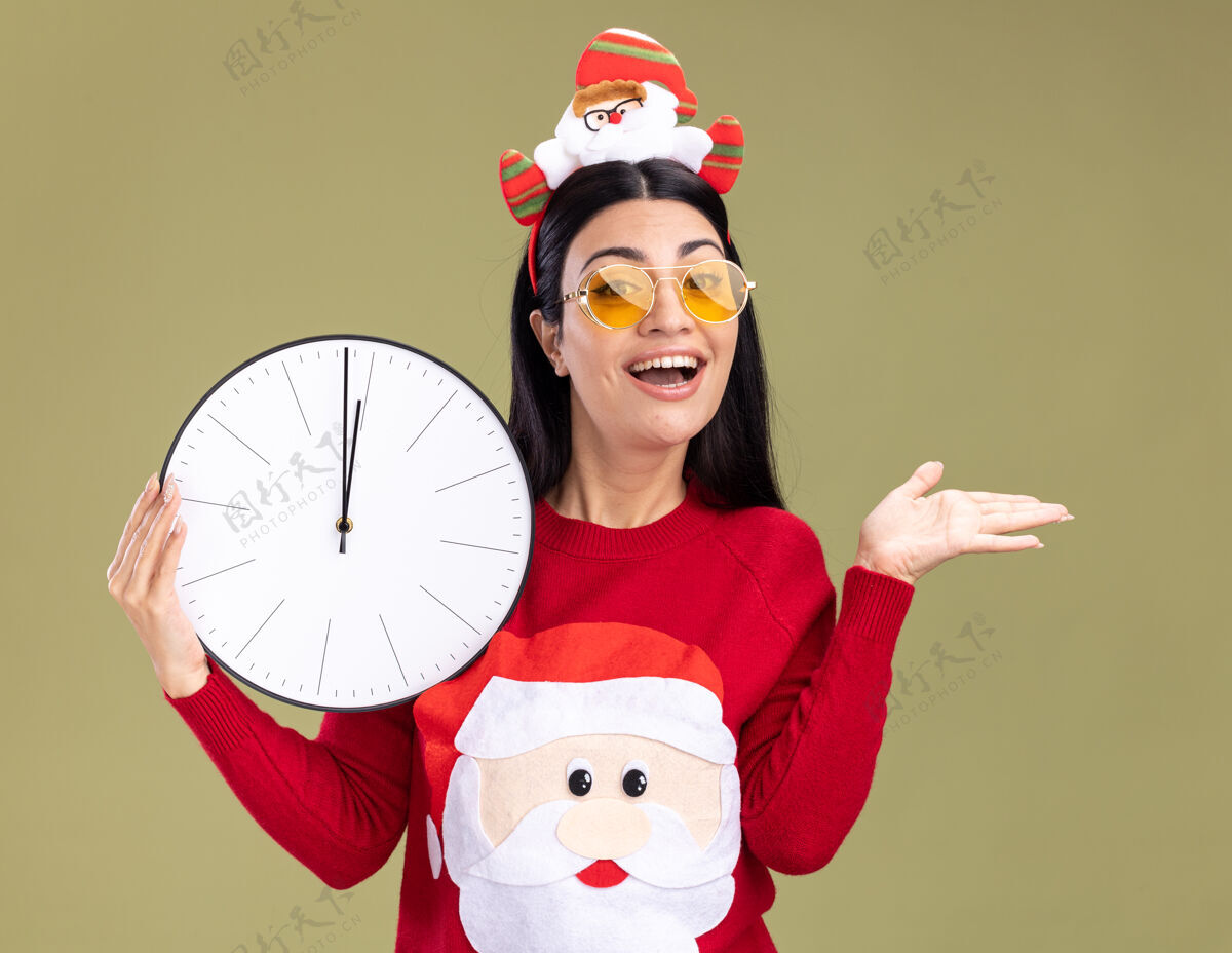 圣诞节快乐的年轻白人女孩戴着圣诞老人的头带和毛衣 戴着眼镜 拿着一只空的手 孤零零地站在橄榄绿的墙上空圣诞老人展示