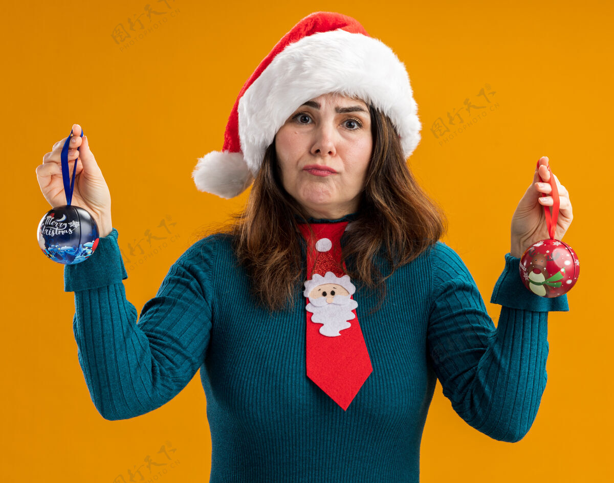球困惑的成年白人妇女 戴着圣诞帽 打着圣诞领带 拿着玻璃球饰物 孤立地放在橙色背景上 留有复制空间圣诞老人橙色领带
