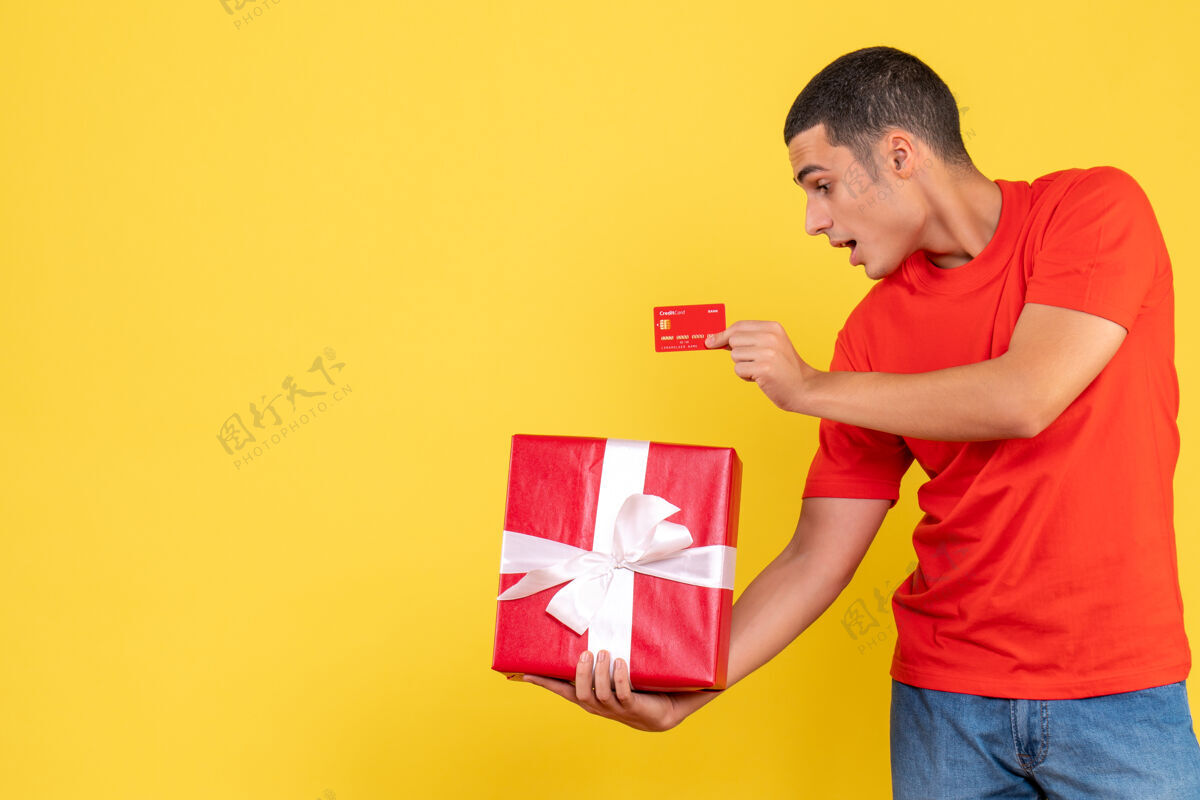 成人年轻人拿着礼物和银行卡在黄色墙上的正面图十二月圣诞节人