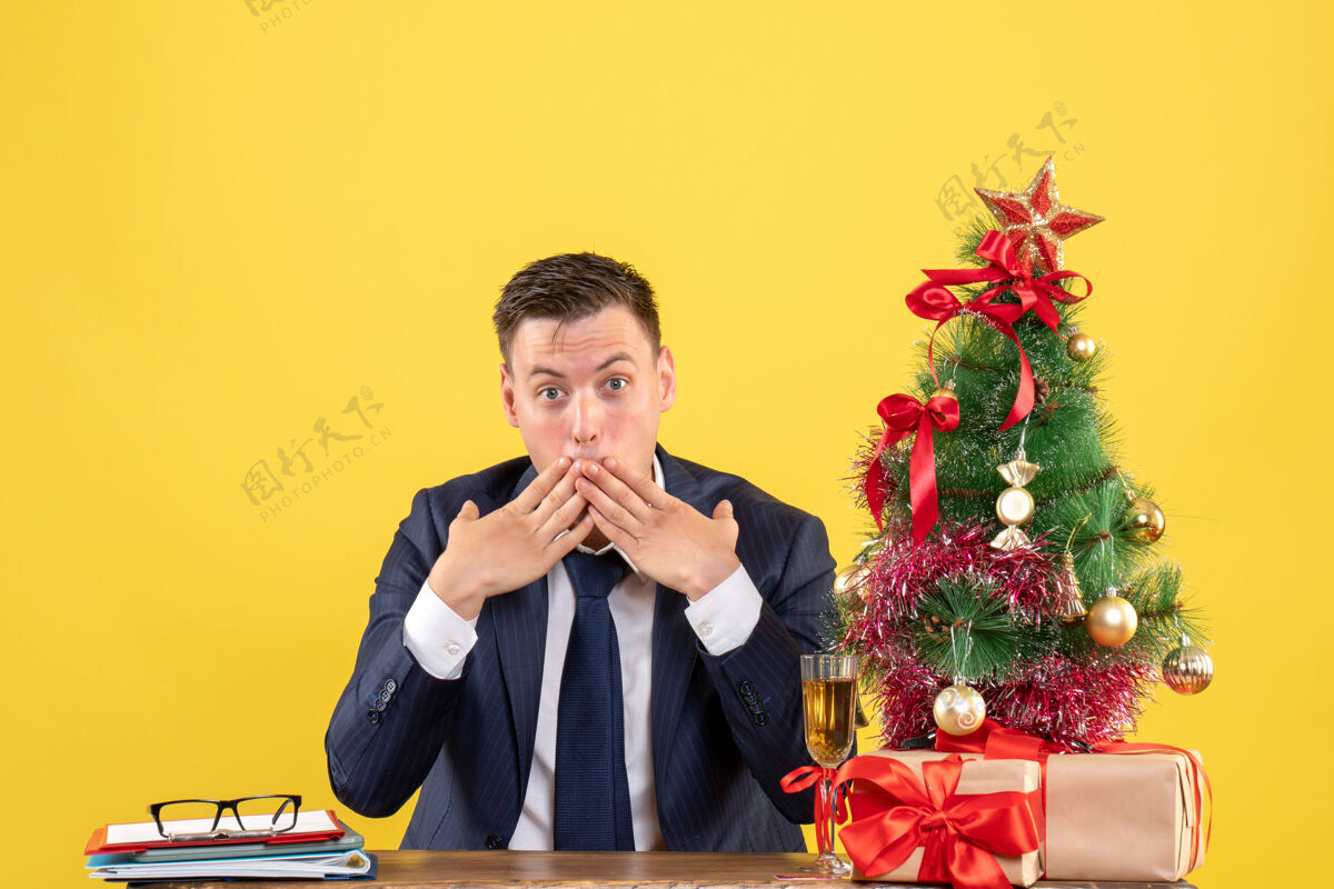 嘴前视图惊讶的人把手放在他的嘴上坐在圣诞树附近的桌子上 黄色的墙上的礼物圣诞节礼物前面