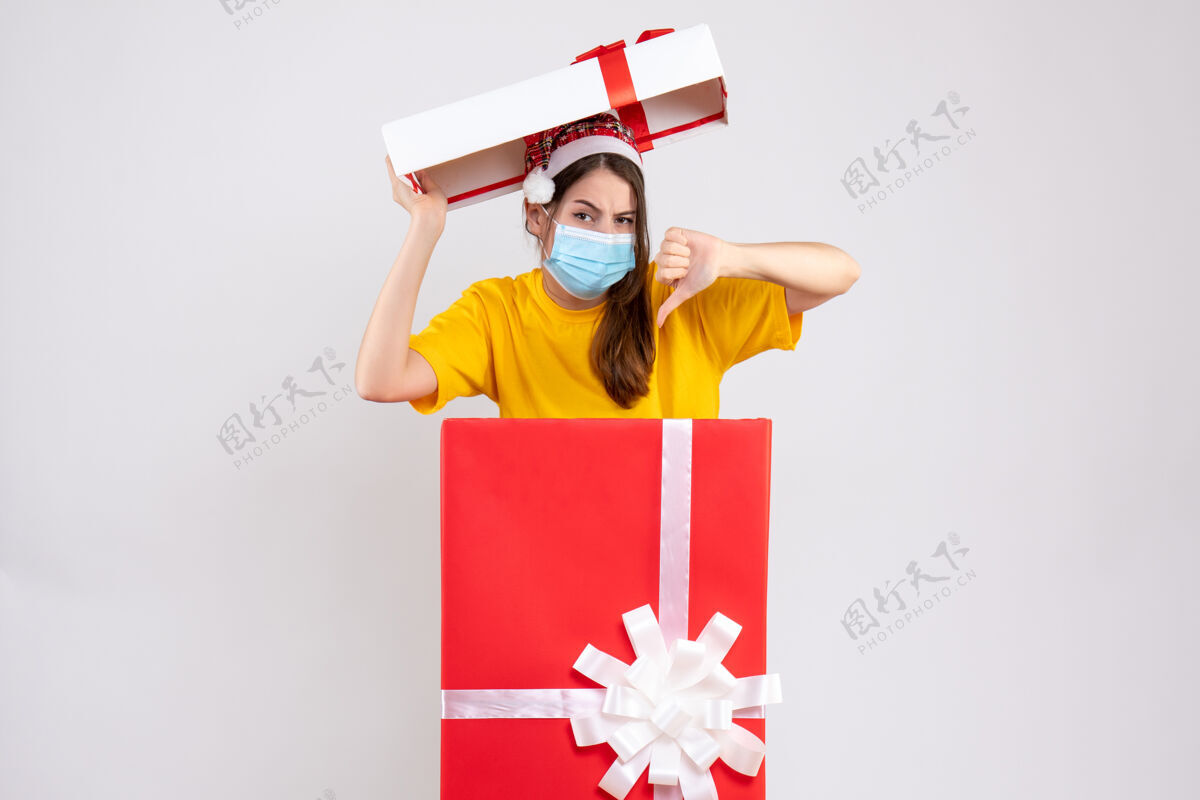 人戴着圣诞帽的快乐女孩站在白色大圣诞礼物后面做拇指朝下的标志礼物漂亮站着