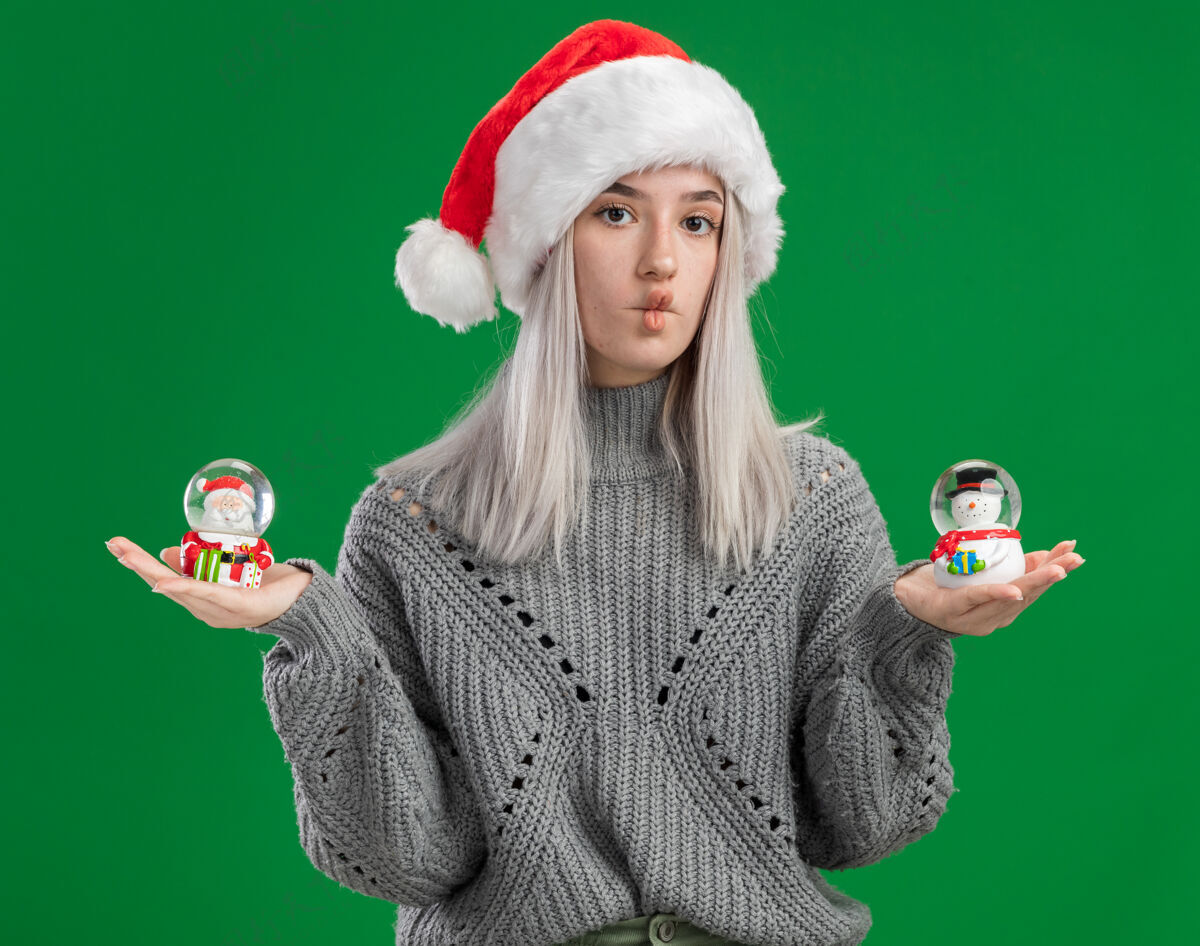 帽子年轻的金发女郎穿着冬衣 戴着圣诞帽 手里拿着圣诞玩具雪球 站在绿色的背景下困惑地看着相机圣诞节圣诞老人雪