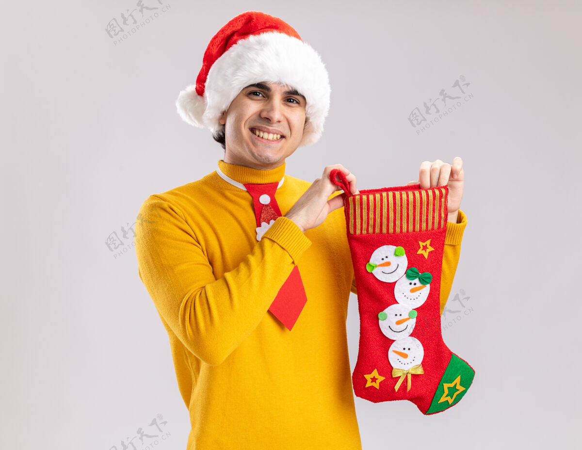 年轻快乐的年轻人穿着黄色高领毛衣 戴着圣诞老人帽 打着滑稽的领带 站在白色的背景上 露出欢快的笑容高领毛衣长袜圣诞节