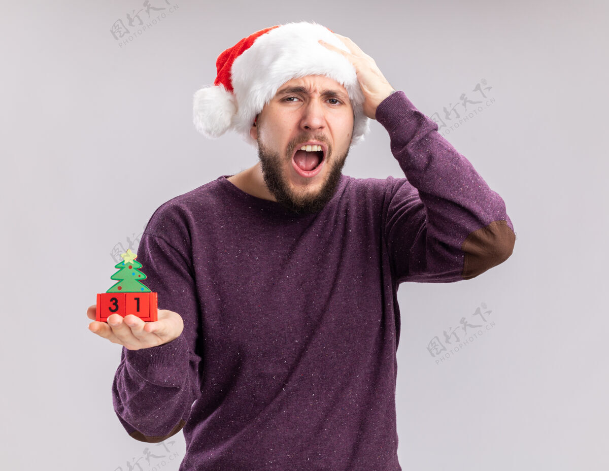 年穿着紫色毛衣 戴着圣诞帽的年轻人站在白色背景上 带着失望的表情高喊着新年约会的方块立方体圣诞老人站着