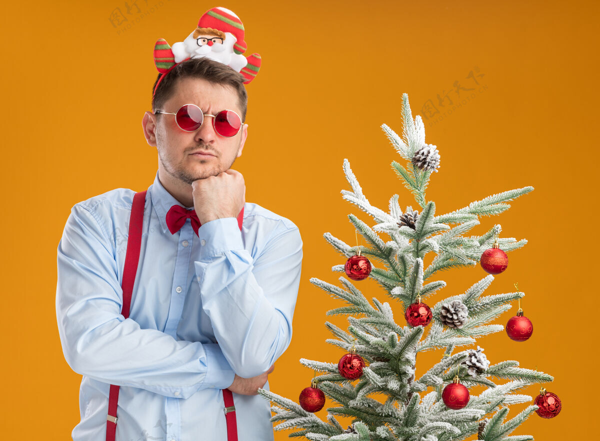 年轻戴着吊带领结的年轻人站在圣诞树旁 戴着圣诞老人和红眼镜 在橙色背景下严肃地看着摄像机站立弓脸
