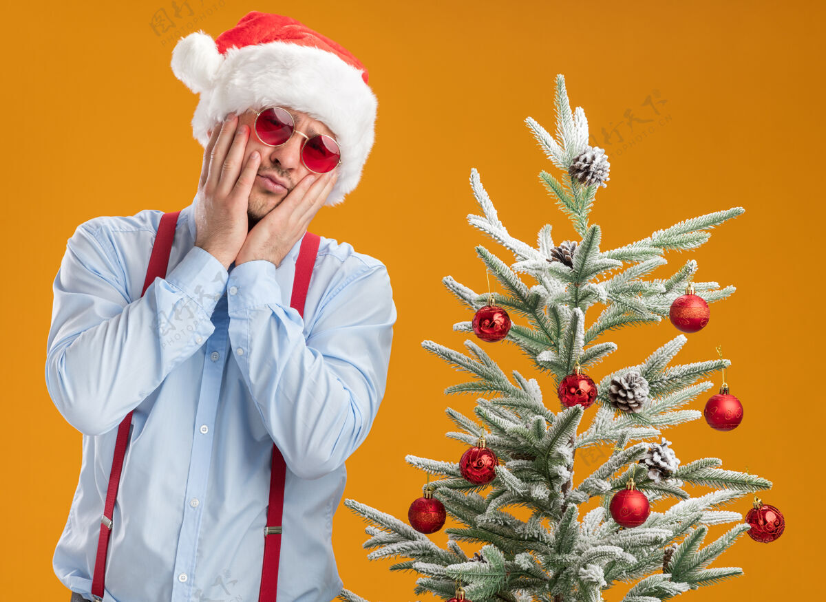 圣诞树一个戴着圣诞帽 戴着红眼镜 系着吊带领结的年轻人站在圣诞树旁 困惑地看着摄像机 在橙色的背景下非常焦虑蝴蝶结吊带帽子