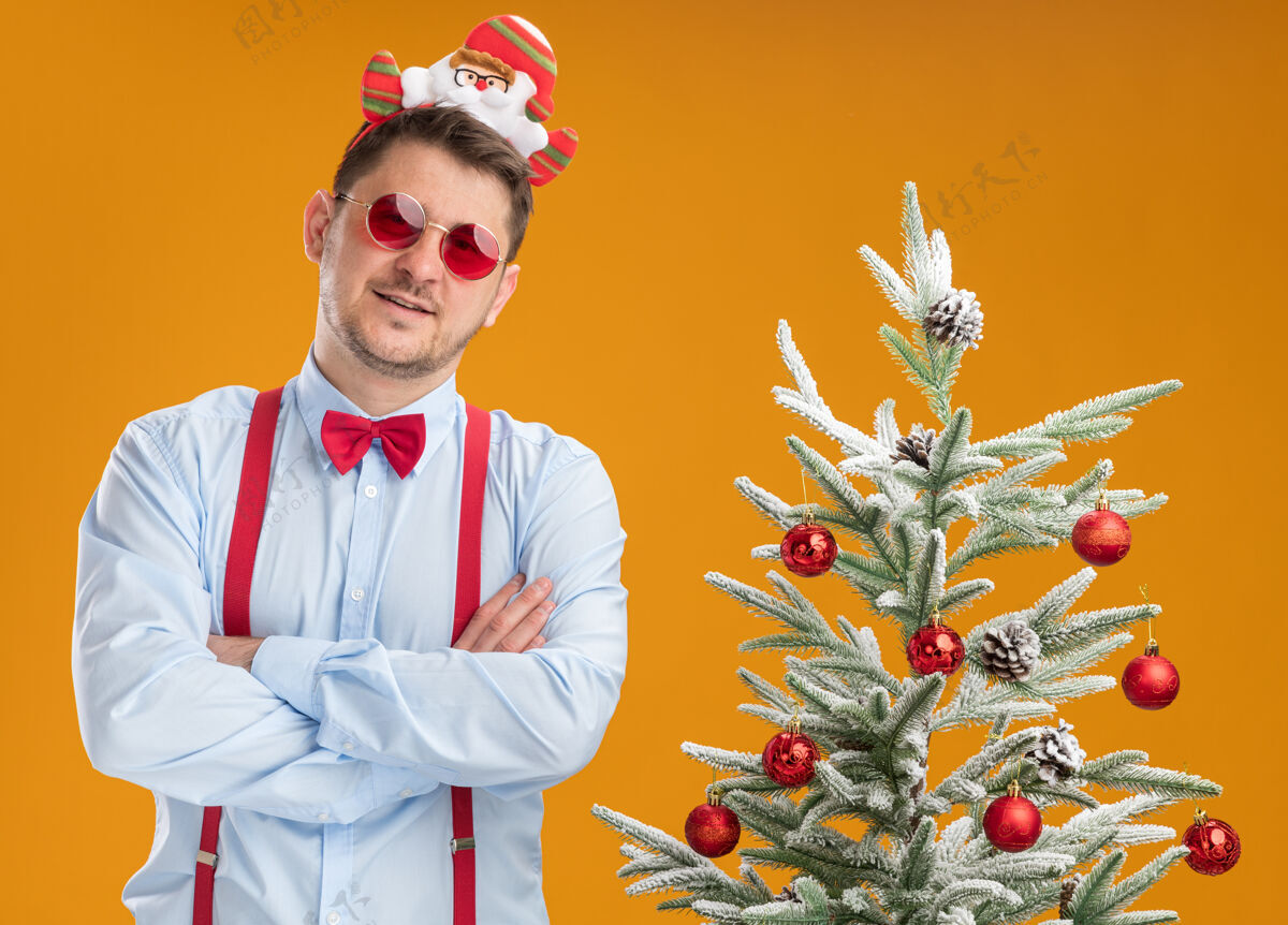 眼镜戴着吊带领结的年轻人站在圣诞树旁 戴着圣诞老人和红眼镜 双臂交叉在橙色背景上 自信地看着摄像机年轻自信蝴蝶结
