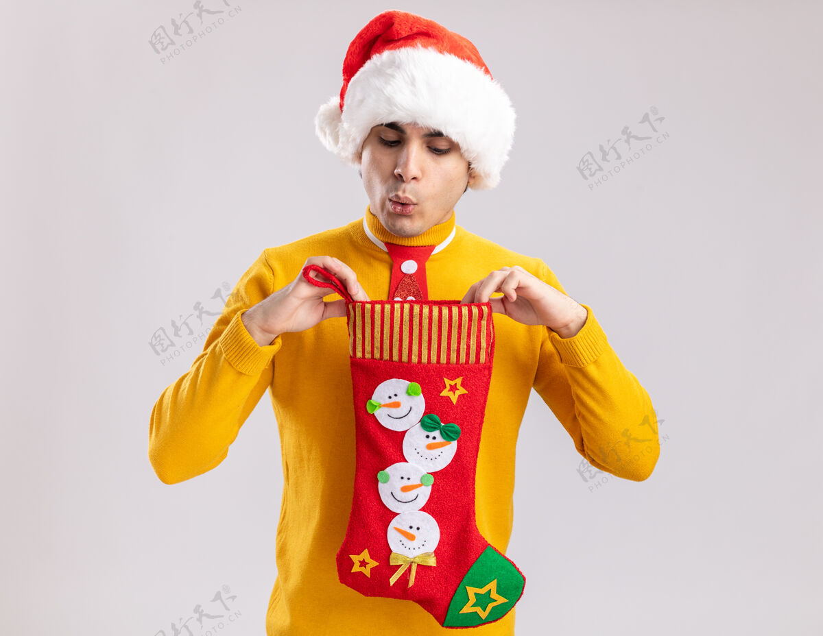 长袜穿着黄色高领毛衣 戴着圣诞老人帽 打着滑稽领带的年轻人拿着圣诞长袜 站在白色背景上看里面圣诞节里面领带
