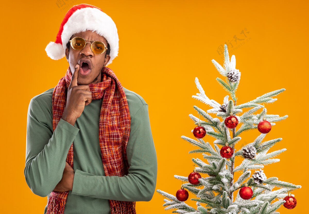 男人一个戴着圣诞帽 脖子上围着围巾的非裔美国人站在橙色墙上的圣诞树旁 惊讶不已惊讶站着到处都是