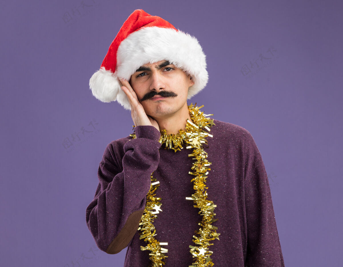 手年轻的胡子男人戴着圣诞老人的帽子 脖子上戴着金属丝 站在紫色的背景上 困惑地看着相机 脸上的手周围金属片站着