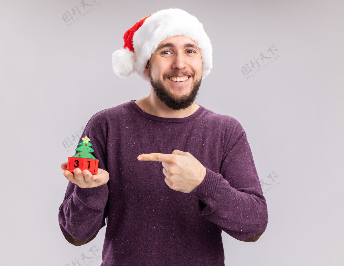 帽子身穿紫色毛衣 头戴圣诞帽的年轻人站在白色背景上 用食指指着方块上的新年日期 快乐而愉快圣诞老人立方体手指