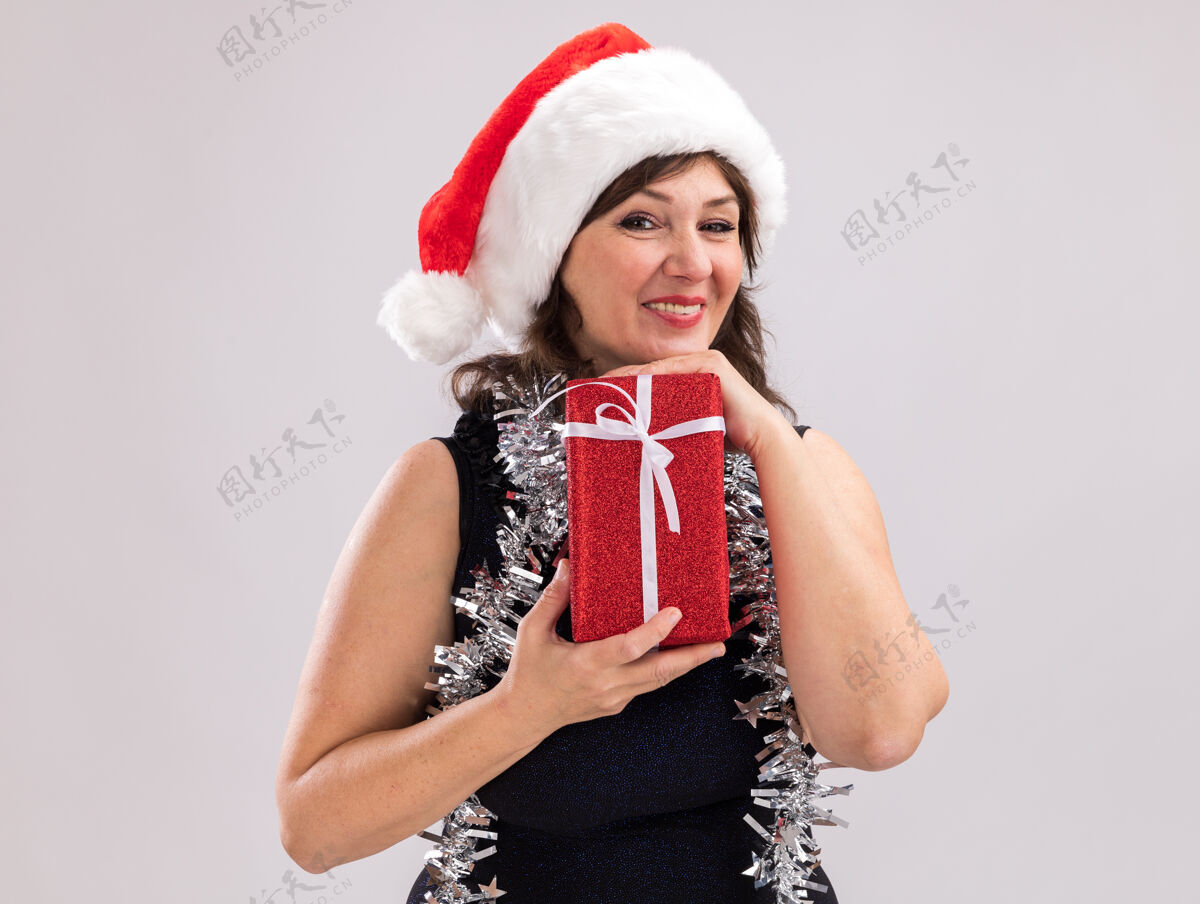 帽子微笑的中年妇女戴着圣诞帽 脖子上戴着金属丝花环 手里拿着圣诞礼包 看着隔离在白色背景上的相机圣诞老人礼物金属丝
