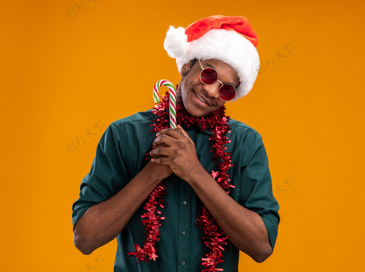 手杖戴着圣诞帽 戴着花环 戴着眼镜 拿着糖果拐杖的非裔美国人站在橙色背景下 面带微笑地看着摄像机拿着圣诞老人帽子