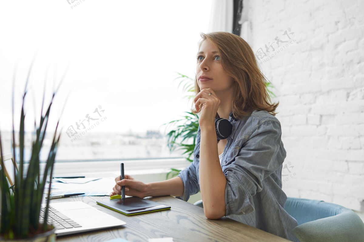 技术一张年轻漂亮的女设计师坐在办公桌前 打开笔记本电脑 拿着笔在绘图板上画画 在家里的办公室里做室内设计项目的照片职业笔艺术家