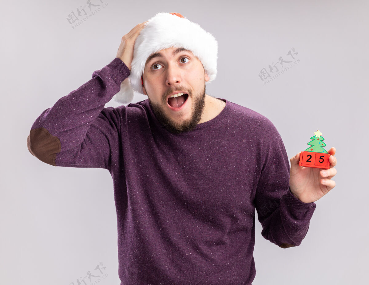 立方体穿着紫色毛衣 戴着圣诞帽的年轻人站在白色的背景上 一边看一边惊奇和惊讶地展示着方块和新年约会帽子惊讶惊讶