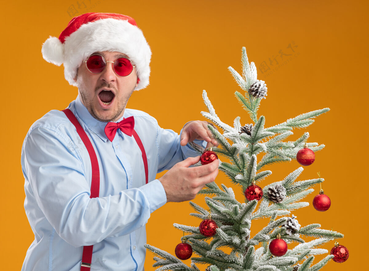 帽子戴着圣诞帽 戴着红眼镜 系着吊带领结的年轻人站在圣诞树旁 把玩具挂在树上 在橙色的背景下又高兴又惊讶年轻悬挂吊带