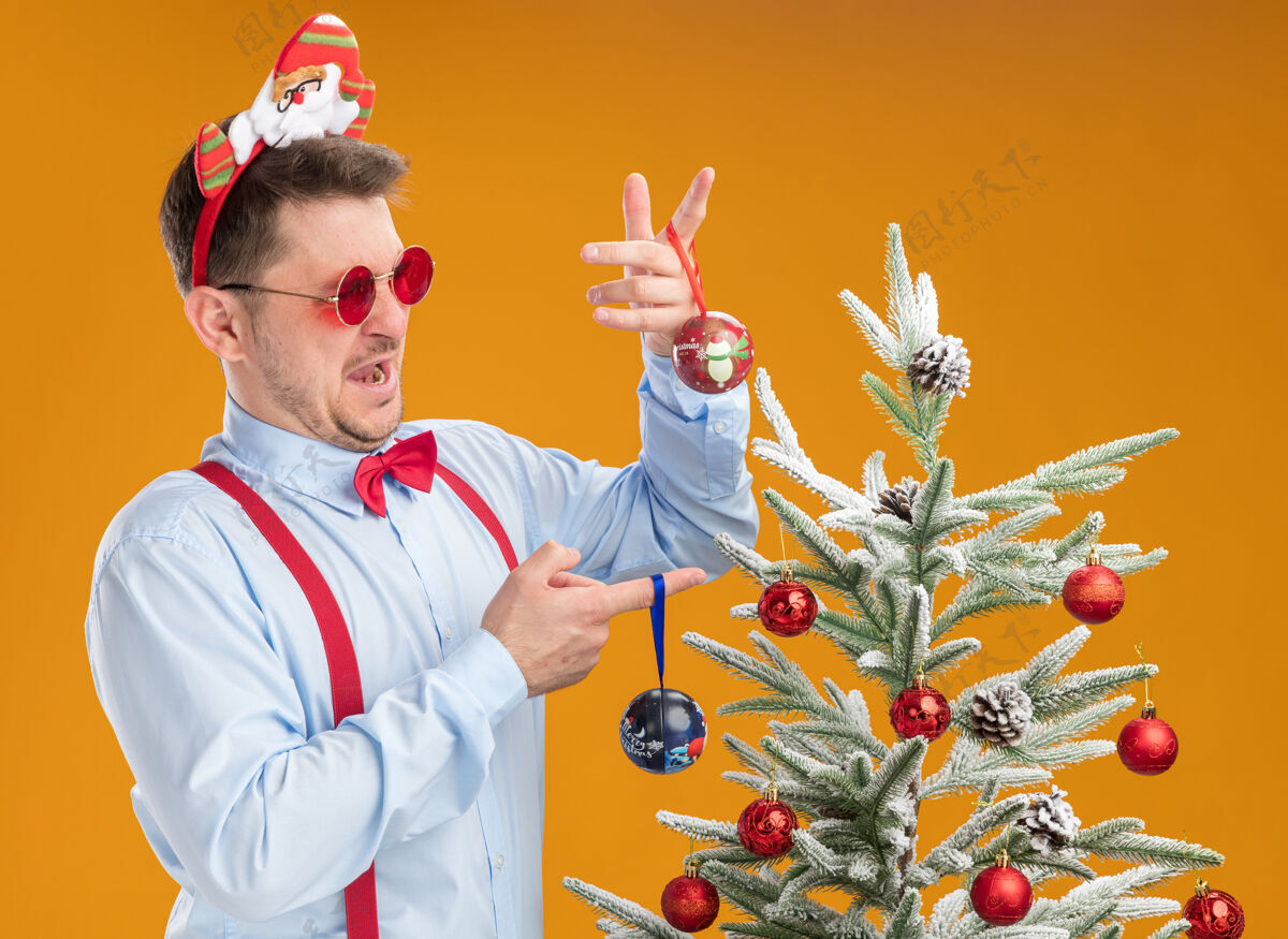 选择戴着吊带领结的年轻人站在圣诞树旁边 戴着圣诞老人和红眼镜 看起来很困惑 试图做出选择 拿着橙色背景的圣诞树玩具蝴蝶结领带圣诞老人