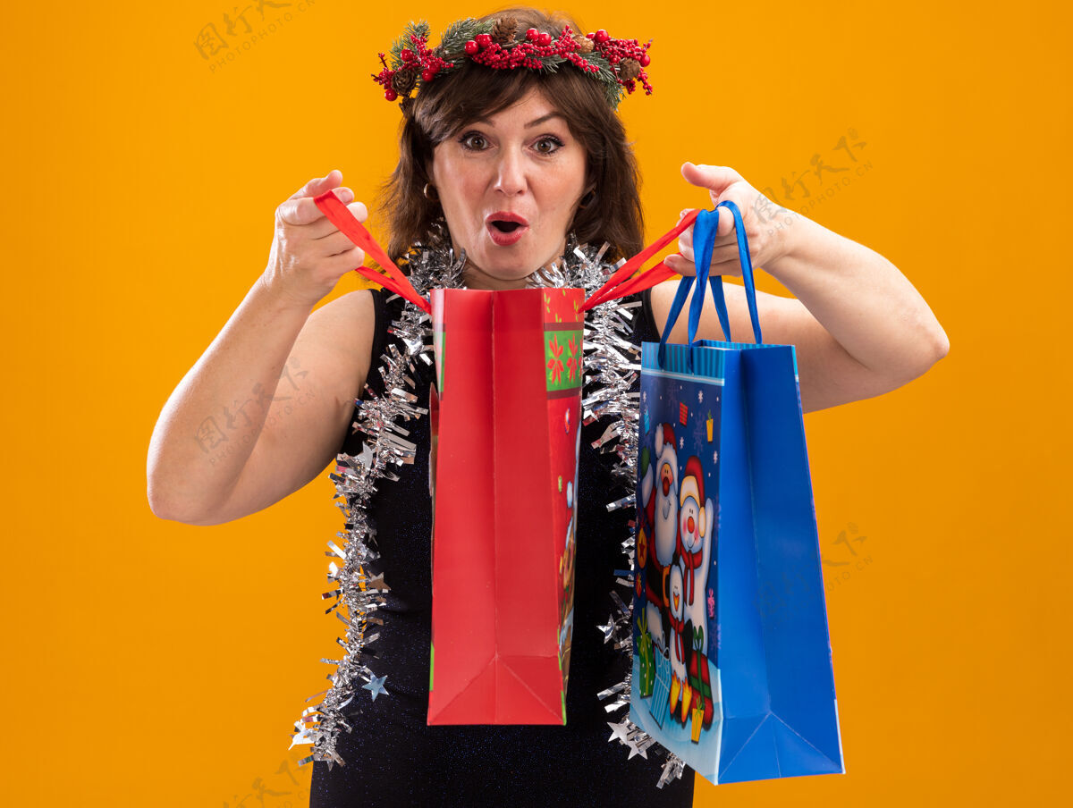 头好奇的中年妇女头戴圣诞花环 脖子上戴着金属丝花环 手里拿着圣诞礼品袋 打开一个 看着孤立在橙色背景上的相机包周围金属丝