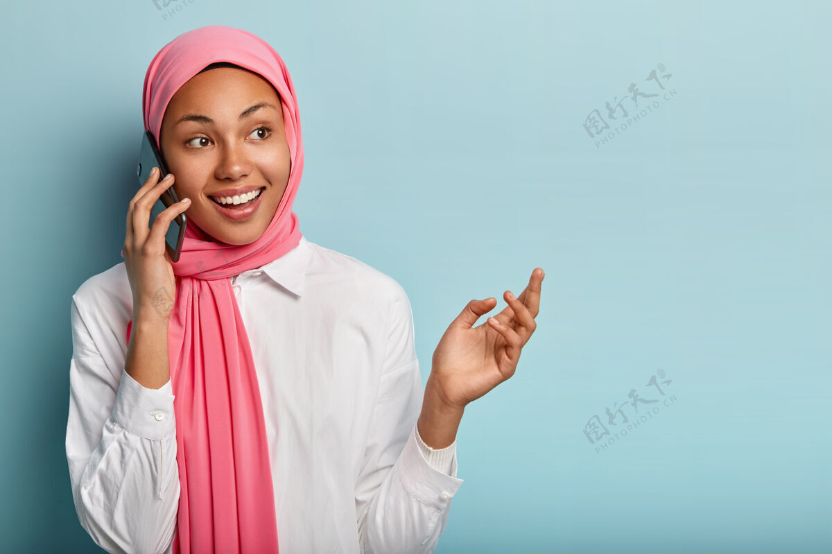打电话开朗的女模特和密友讨论好消息 把智能手机放在耳边 看起来很开心墙壁抱着穆斯林