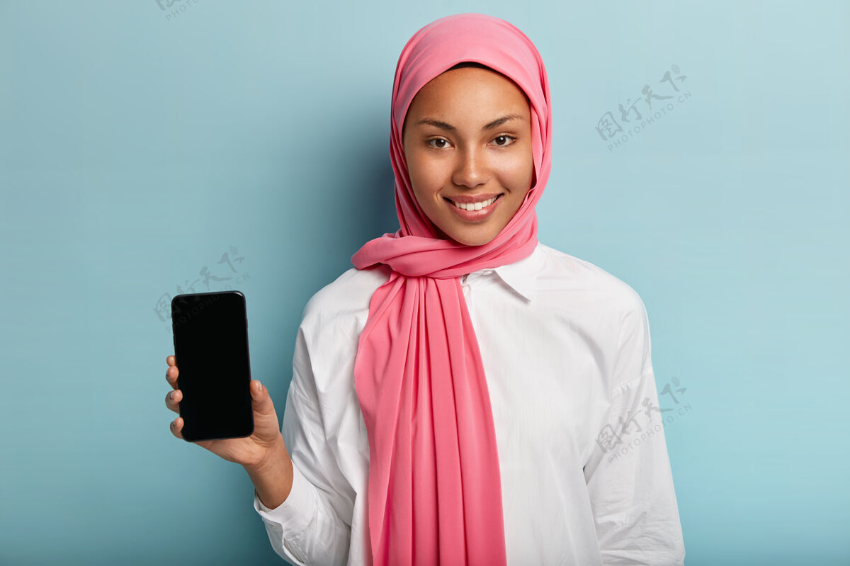 阿拉伯美丽的穆斯林妇女为你的广告做现代小玩意广告 手持白屏智能手机装置 头戴传统面纱民族电话乐观