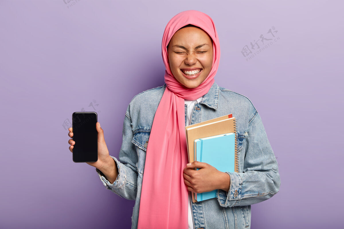 设备这是你需要的手机有伊斯兰观点的快乐女人 戴着传统的头巾 展示智能手机屏幕 大笑手机日记大笑