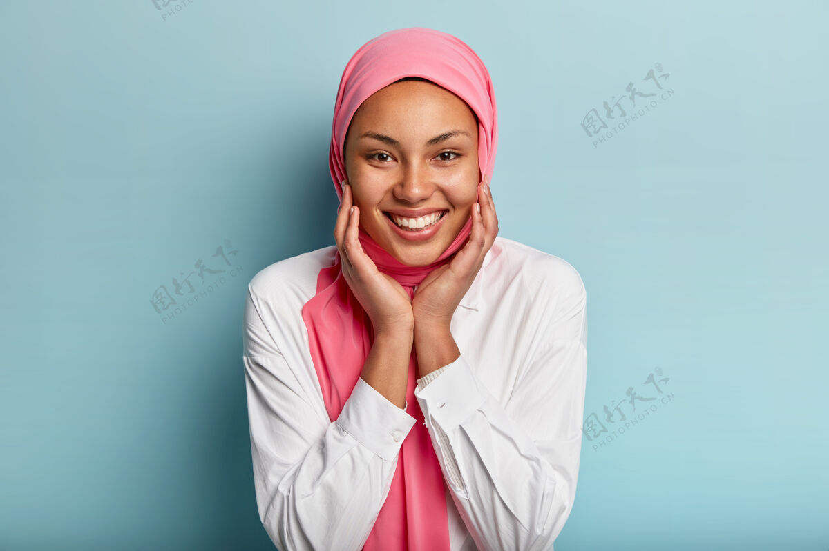 表情面容姣好的穆斯林妇女的头像用双手抚摸脸颊 露出洁白的牙齿 穿着白衬衫和粉色面纱 与蓝色的墙壁隔离 表达喜悦 幸福 喜悦室内习俗妇女