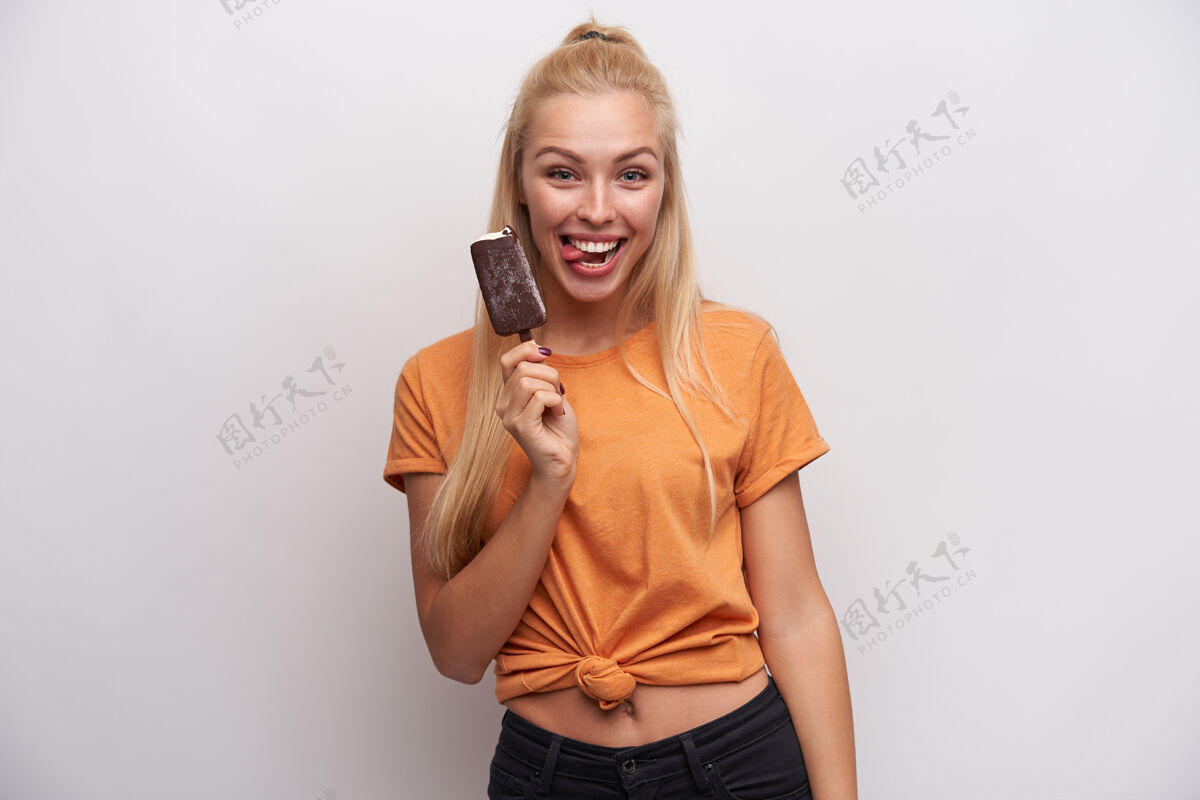 长有趣的照片 快乐的年轻长发金发女子在橙色t恤展示愉快的舌头对相机 而在白色的背景与冰淇淋摆姿势棍棒20多岁女性