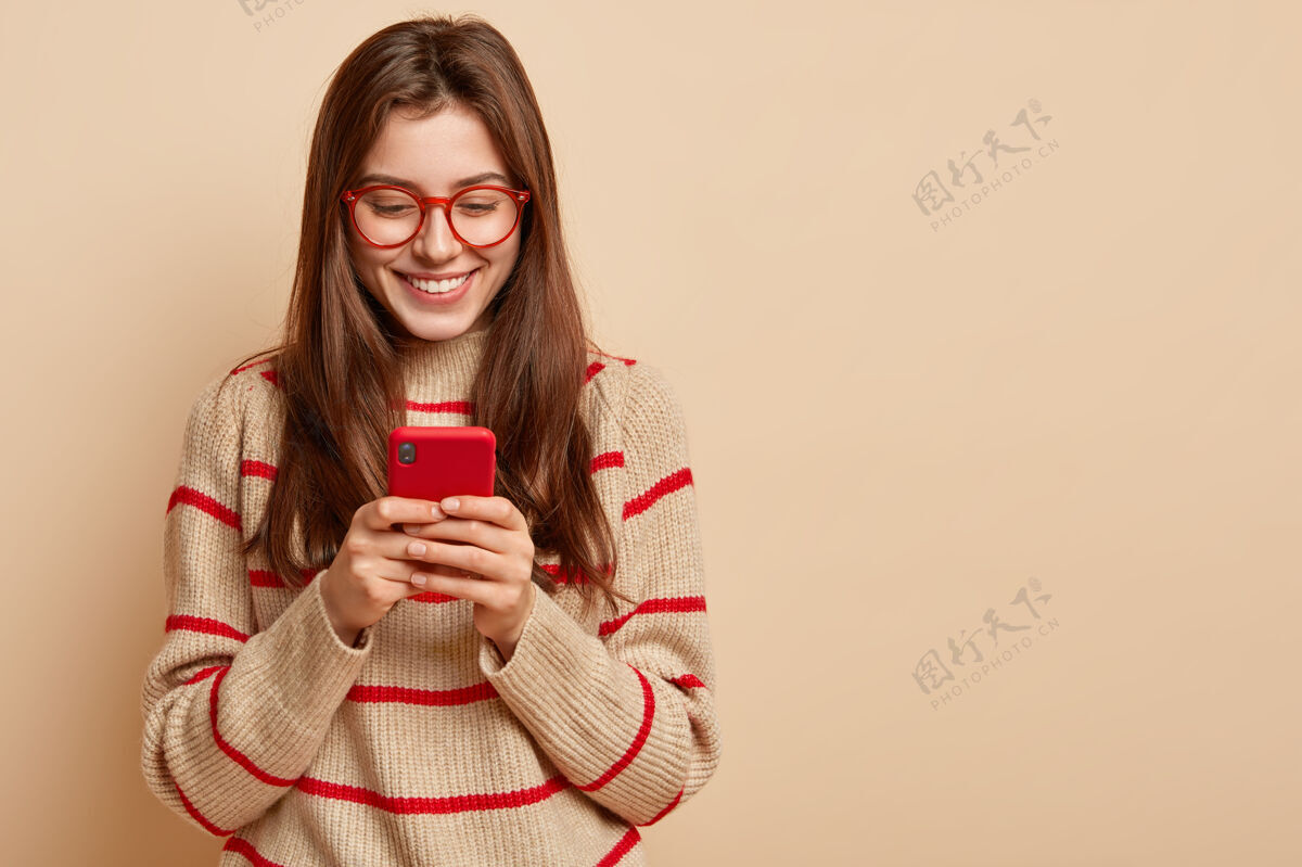 阅读满足少女的室内照片在手机上发短信 在线阅读有趣的文章 穿着休闲装 在自己的网页上创建新的出版物 在棕色的墙壁上与自由空间隔离应用程序手机肖像