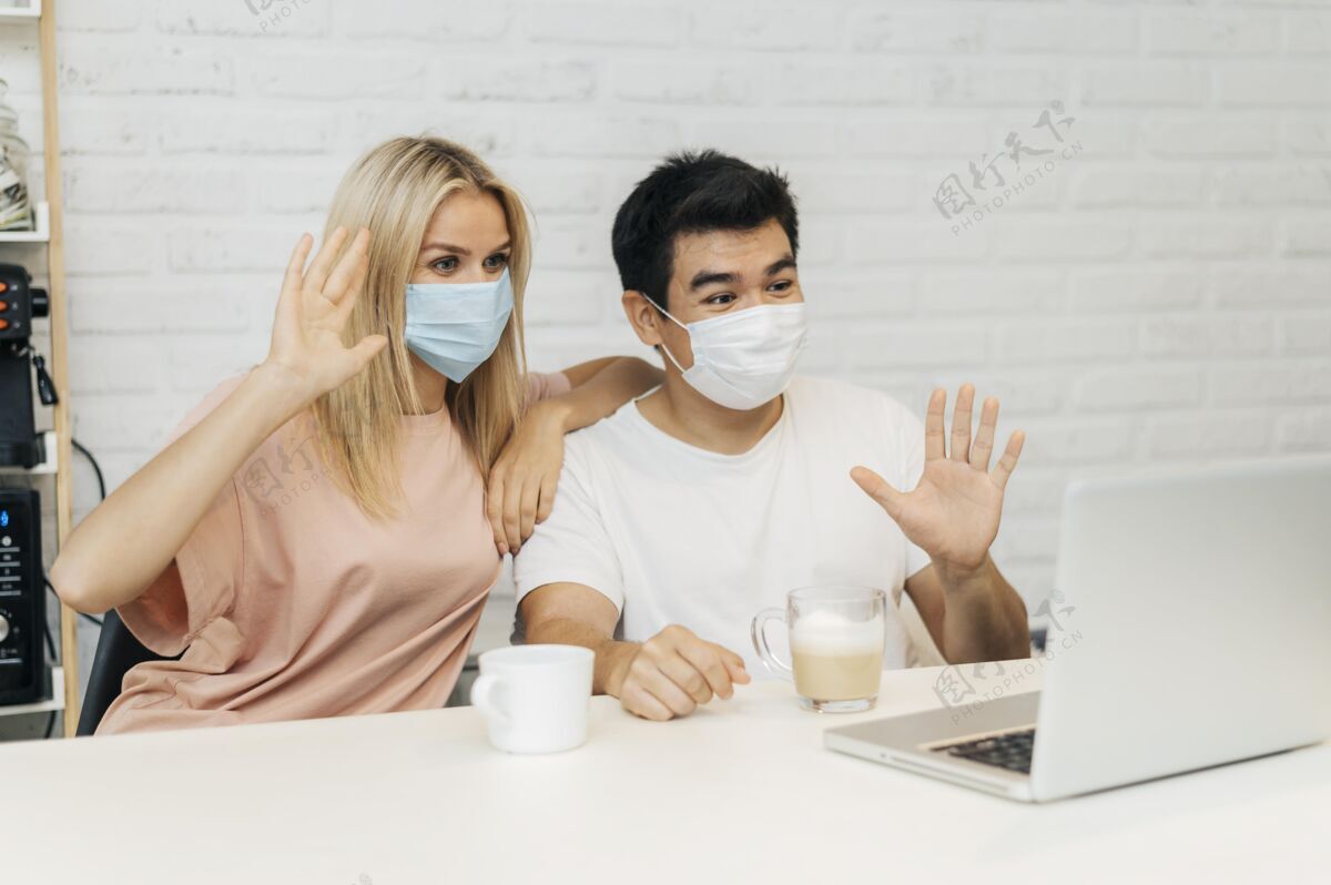 距离流感大流行期间 一对戴着医用口罩的夫妇在家里对着笔记本电脑挥手病毒杯子Covid19