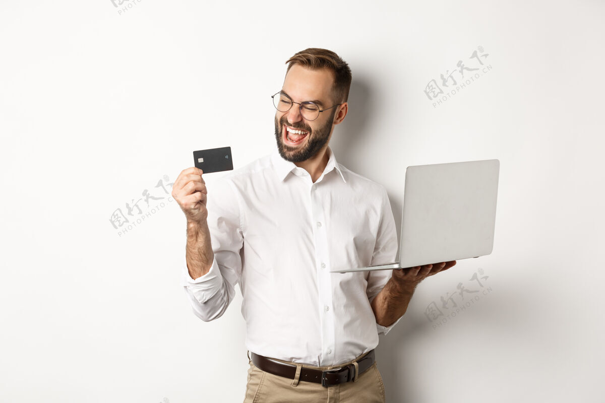 商人网上购物满意的帅哥看着信用卡下单后上网 用笔记本电脑 站着信用卡胡须员工