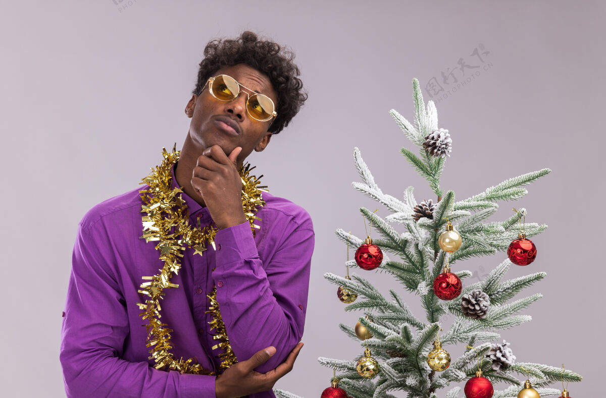 眼镜体贴的美国黑人年轻人戴着眼镜 脖子上戴着金箔花环 站在装饰好的圣诞树旁 手放在下巴上 在白色的背景下孤立地抬头看金属片保持花环