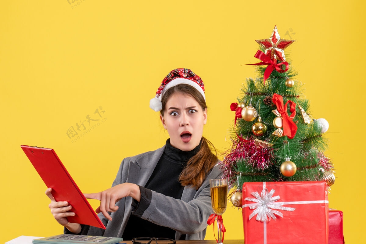 圣诞节前视图困惑的女孩戴着圣诞帽坐在桌边文档文件夹圣诞树和礼物鸡尾酒在桌上黄色的背景文件夹快乐女孩