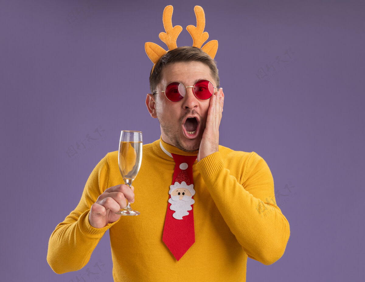 高领毛衣一个穿着黄色高领毛衣 戴着红色眼镜的年轻人 打着有趣的红色领带 戴着鹿角边 手里拿着一杯香槟 站在紫色的背景下 惊讶而又惊奇地看着镜头年轻领带香槟