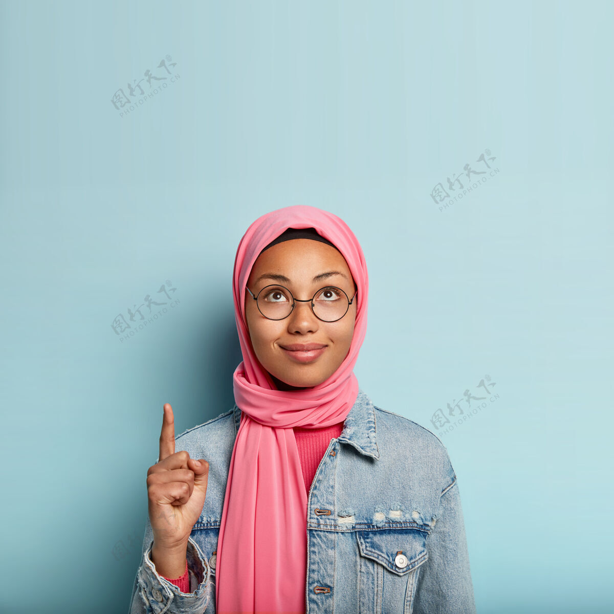 夹克迷人的年轻女子 皮肤黝黑健康 面朝上 用食指指指 头戴粉色头巾 身穿时尚夹克 站在蓝色墙壁上 广告复制空间 展示正确的方式Indexfinger向上上衣